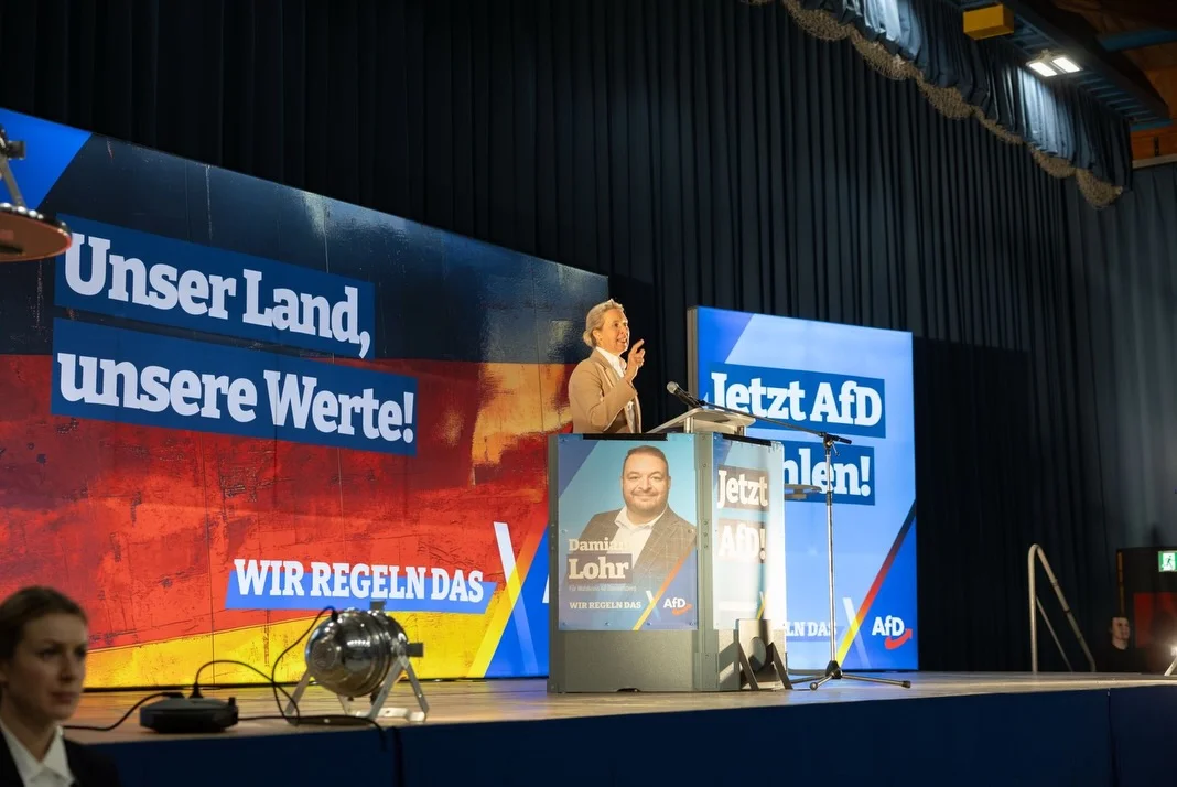 Photo by Alice Weidel on February 21, 2026. May be a video game screenshot of television, speaker, microphone, screen, dais and text that says 'Unser UnserLand, Land, unsere Werte! letztAfD Jetzt hlen! AM! WIR REGELNDAS Damin Lohr WIR VIRREGELNDAS EGELN AFD INDA: AFD'.